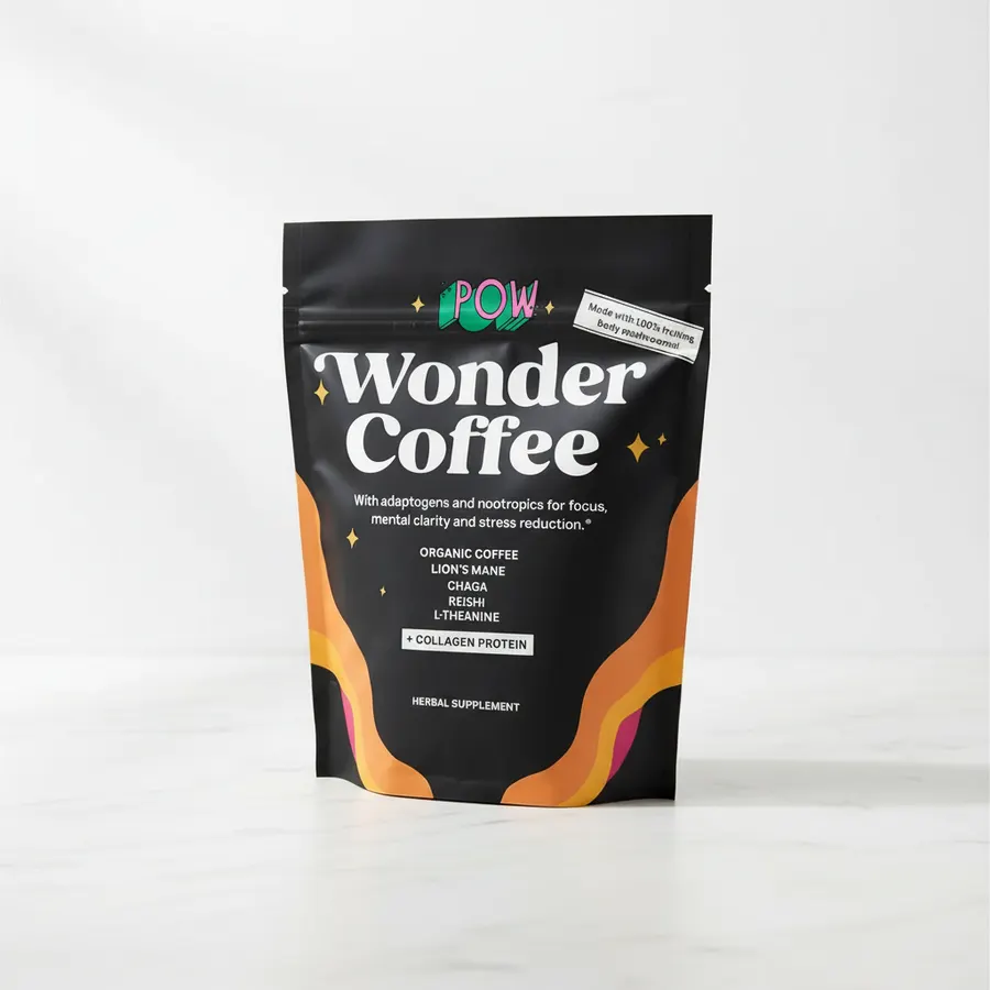 A woman over 50 receiving a small coffee packet from a friend's hand across a kitchen counter, both smiling warmly. Mornin...