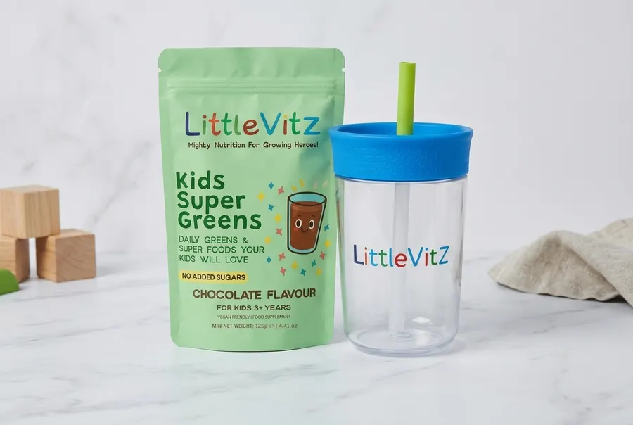 A frosted glass jar of children's greens powder supplement with a filled scoop resting beside it on a bright breakfast tab...
