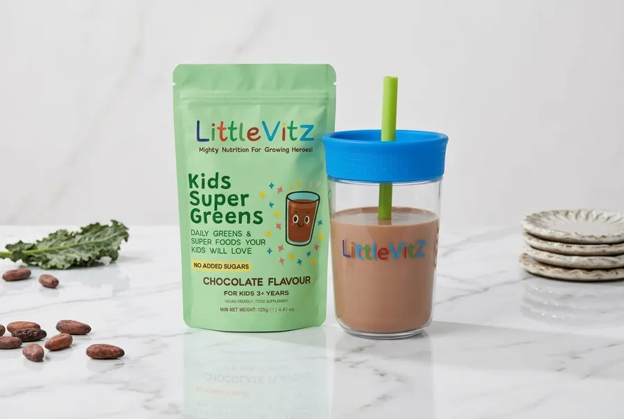A matte pouch of chocolate-flavoured kids greens powder open on a breakfast table beside a scoop of fine powder, a glass o...