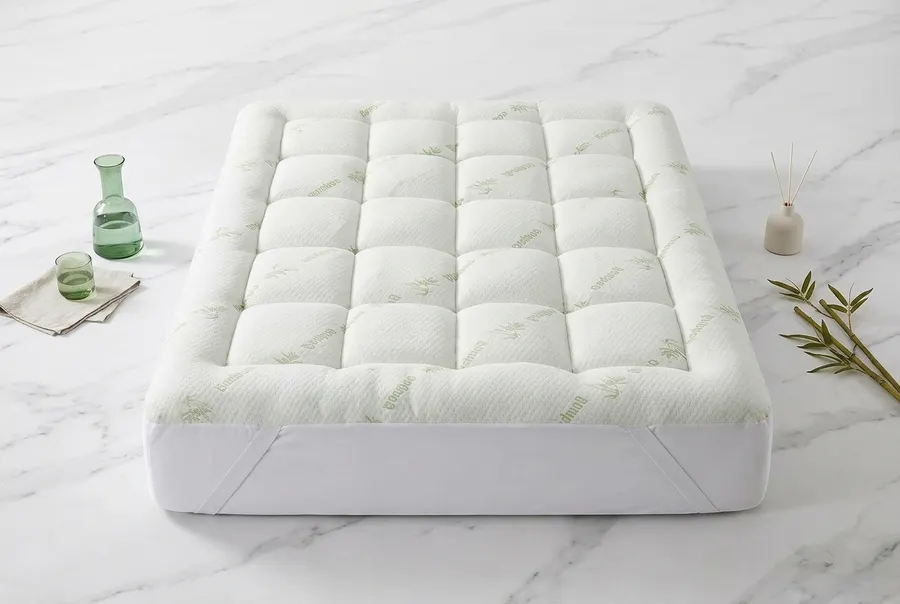 A plush white organic bamboo mattress topper positioned at a slight angle on a neatly made bed in a warm, sunlit bedroom. ...