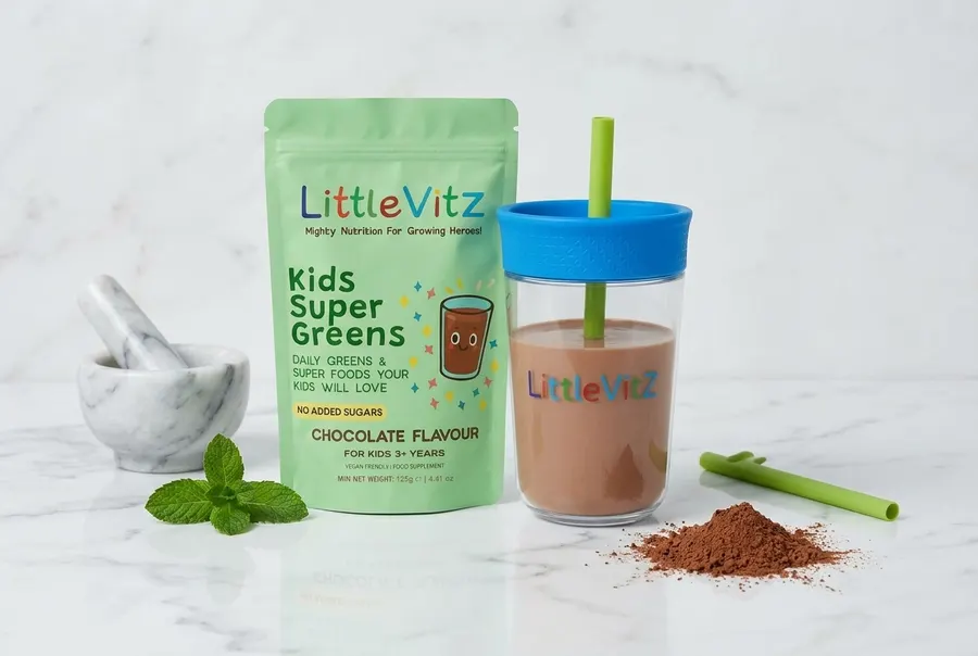 A frosted glass bottle of green superfood powder sits upright on a light wood kitchen counter next to a clear glass filled...
