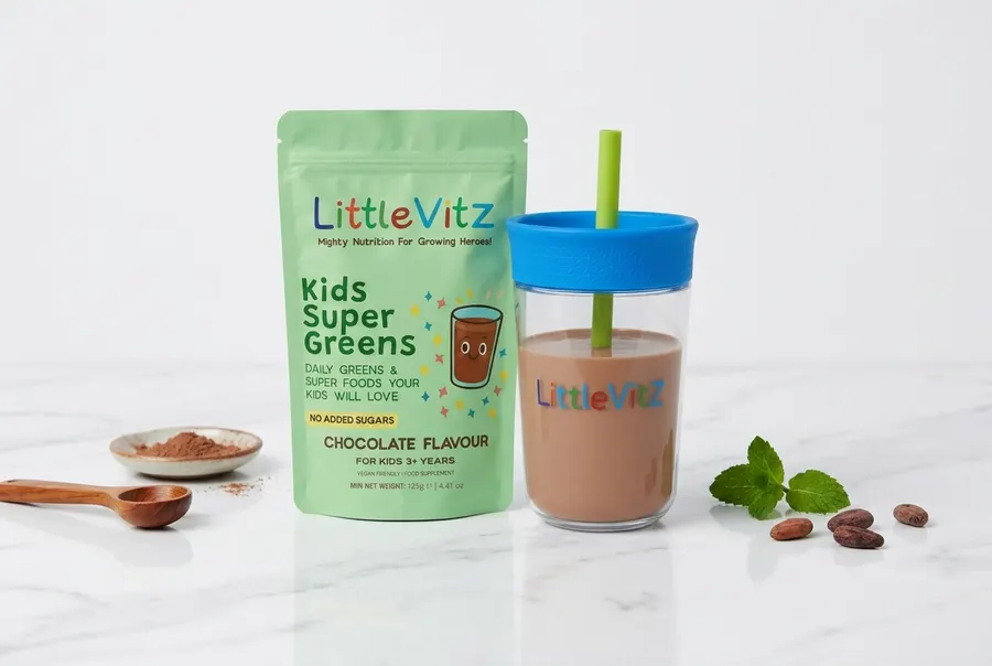 A frosted white plastic tub of children's greens powder next to a tall glass of rich chocolate-brown smoothie on a bright ...
