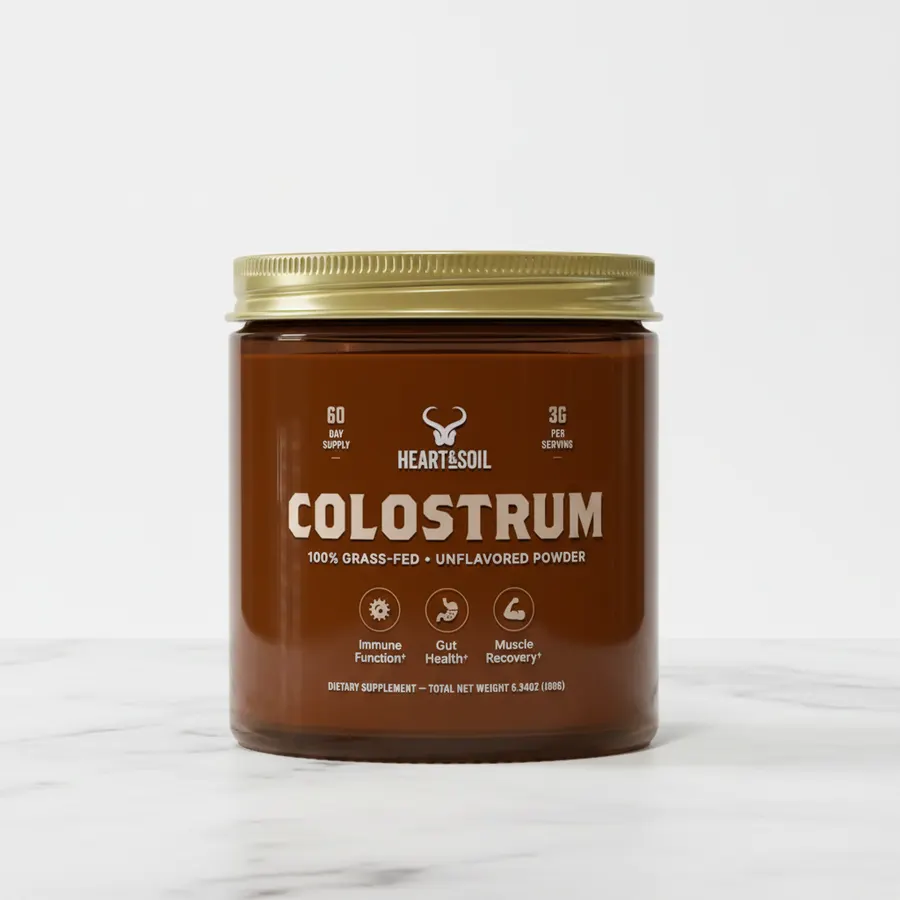Close-up of a frosted glass jar of grass-fed colostrum powder with a wooden spoon resting in it, surrounded by fresh pastu...