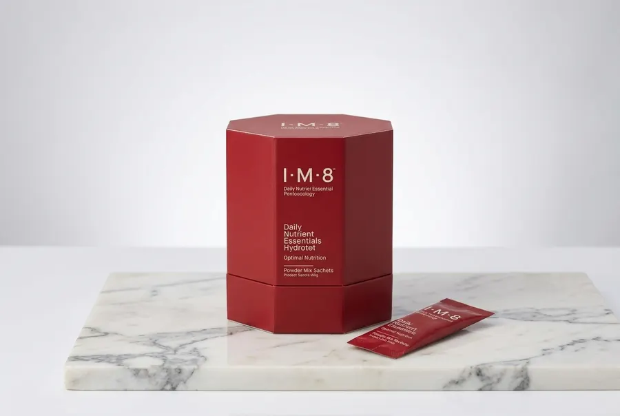 A matte canister of daily greens powder centered upright on a clean marble countertop with a filled glass of green drink b...