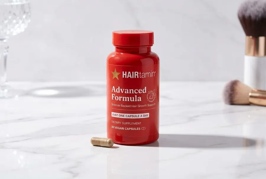 A frosted white capsule bottle of biotin hair supplement stands upright on a clean two-tone seamless backdrop split horizo...