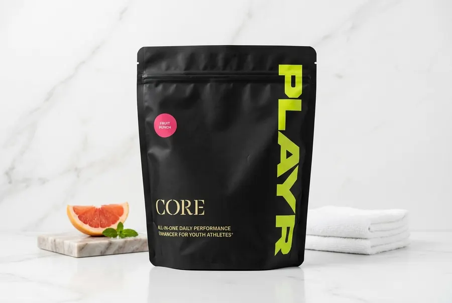 A frosted white tub of youth sports recovery supplement on a clean kitchen counter next to a glass of water and a soccer b...
