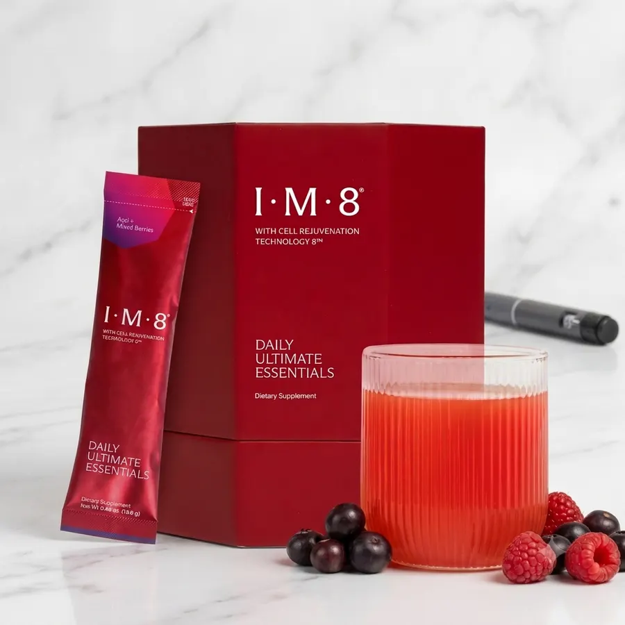 A single slim matte sachet of multivitamin powder beside a clear glass of mixed amber-tinted drink on a clean white marble...