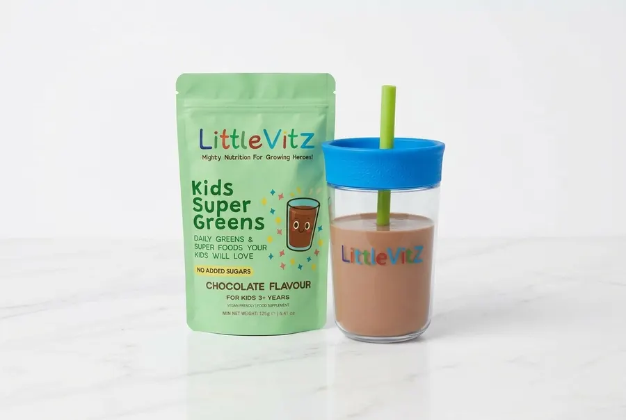 A frosted glass jar of chocolate greens powder for children on a clean white marble kitchen counter, next to a glass of ch...