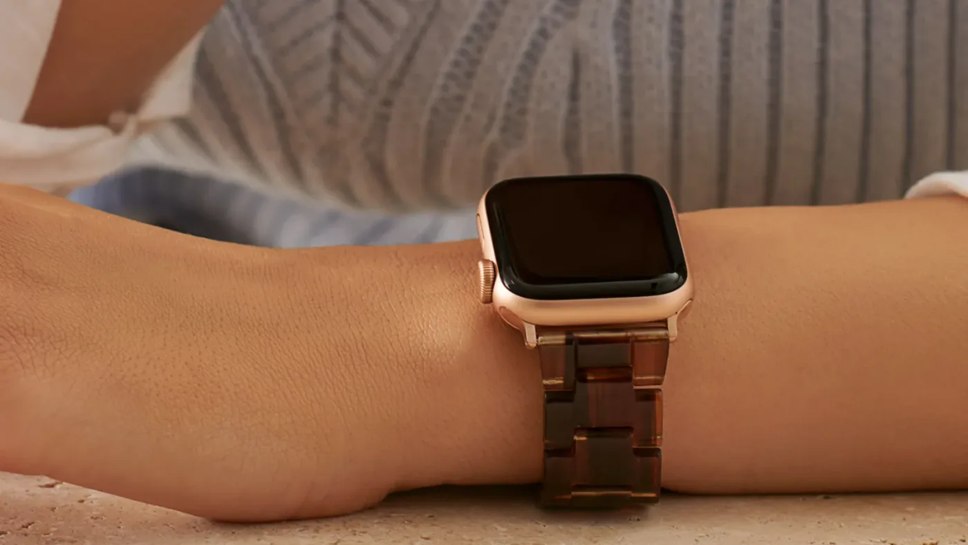 A frosted stainless steel woven link Apple Watch band resting diagonally across a clean white surface, the band's twisted ...