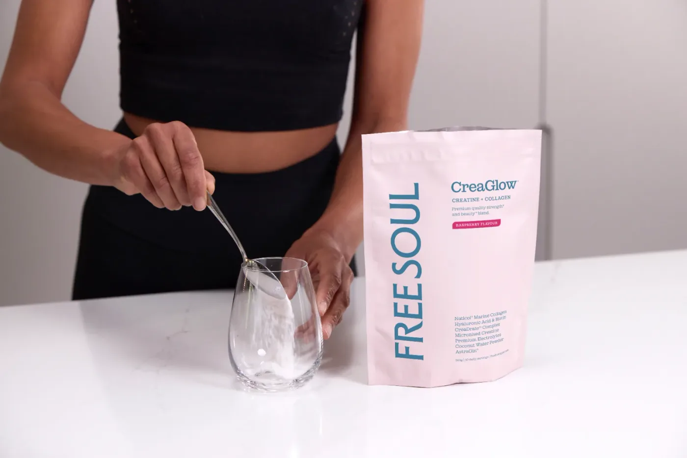 A frosted pink tub of powdered supplement with a clean matte label, centered on a pale marble countertop beside a filled g...
